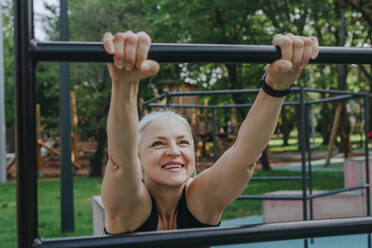Happy mature woman stretching on bars in playground - YTF00046