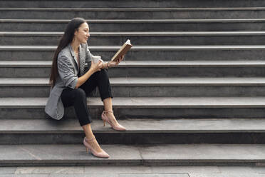 Lächelnde Geschäftsfrau, die ein Buch liest und mit einem Kaffeebecher auf einer Treppe sitzt - VPIF07114