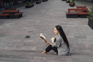Businesswoman with coffee mug reading book on steps at office park - VPIF07115