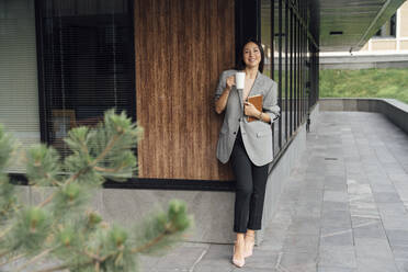 Lächelnde Geschäftsfrau mit Kaffeebecher und Buch vor einem Bürogebäude stehend - VPIF07116