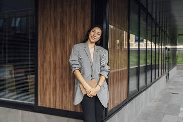 Lächelnde nachdenkliche Geschäftsfrau mit Buch vor einem Bürogebäude stehend - VPIF07118