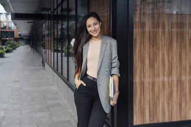 Smiling businesswoman with hand in pocket standing outside office building - VPIF07120