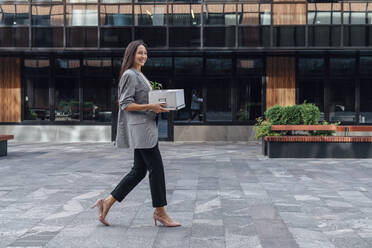 Smiling businesswoman walking with box in front of office building - VPIF07140