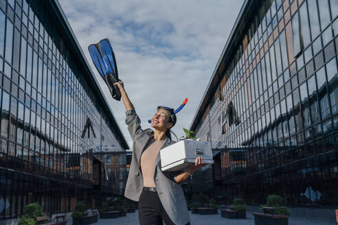 Glückliche Geschäftsfrau mit Tauchflosse und Box vor einem Bürogebäude - VPIF07142