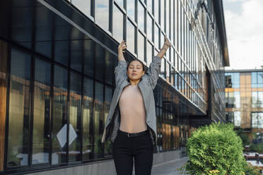 Beautiful businesswoman with eyes closed stretching arms in front of office building - VPIF07160