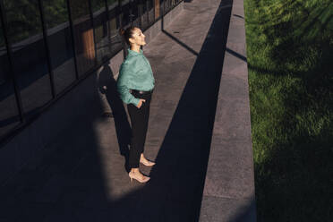 Lächelnde Geschäftsfrau mit Hand in der Tasche genießt das Sonnenlicht im Büropark - VPIF07174