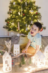 Fröhlicher Junge mit Katze spielt in der Nähe von Weihnachtsbaum zu Hause - ONAF00108