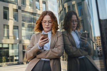 Geschäftsfrau mit Brille, die ein Smartphone benutzt und sich an eine Glaswand lehnt - VPIF07231