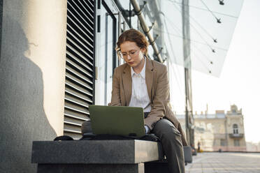 Junge Geschäftsfrau mit Laptop auf einer Bank neben einem Bürogebäude - VPIF07247