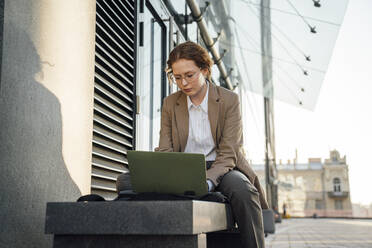 Young businesswoman using laptop on bench by office building - VPIF07247