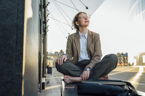 Young businesswoman with eyes closed sitting cross-legged by office building - VPIF07252