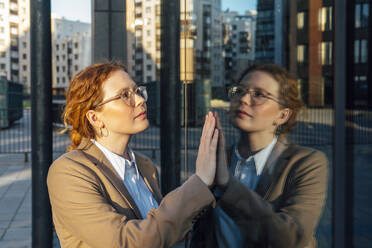 Thoughtful young redhead businesswoman touching reflection on glass wall - VPIF07259