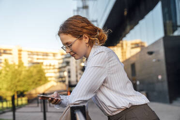 Junge rothaarige Geschäftsfrau, die ein Mobiltelefon benutzt und sich auf ein Geländer stützt - VPIF07277