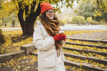 Happy young woman holding disposable cup by staircase at park - OYF00746