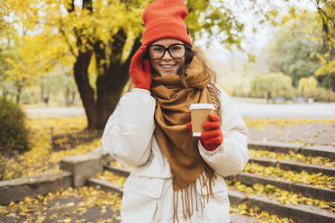 Happy woman wearing eyeglasses holding disposable cup at park - OYF00747