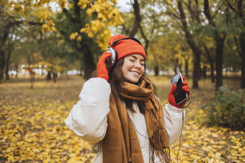 Glückliche junge Frau mit Handy in der Hand, die im Park Musik über Kopfhörer hört - OYF00754