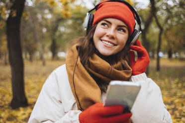 Lächelnde junge Frau mit Handy in der Hand, die im Park über Kopfhörer Musik hört - OYF00761