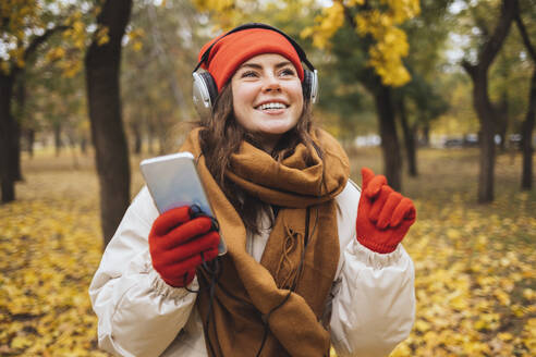 Glückliche Frau mit Mobiltelefon, die im Park Musik über Kopfhörer hört - OYF00762