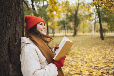 Thoughtful smiling woman with book leaning on tree trunk at park - OYF00766