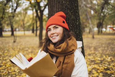 Thoughtful smiling young woman with book in front of tree trunk at park - OYF00767