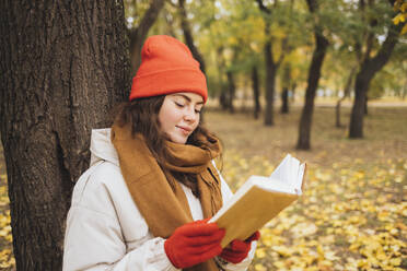 Smiling woman reading book by tree trunk at park - OYF00768