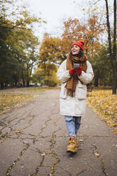 Glückliche junge Frau mit Smartphone in der Hand, die auf einem Fußweg im Park spazieren geht - OYF00772