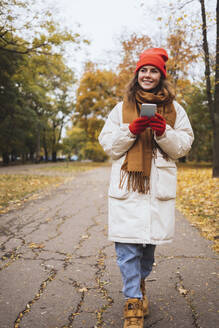 Smiling young woman with mobile phone walking on footpath at park - OYF00773