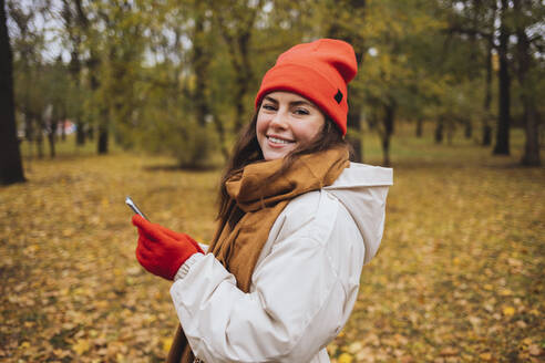 Smiling woman wearing knit hat holding mobile phone in autumn park - OYF00775