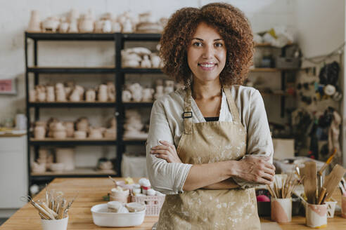 Smiling potter wearing apron standing with arms crossed in workshop - YTF00087