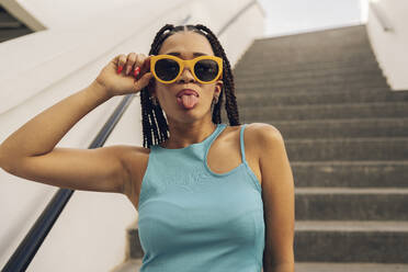 Young woman wearing sunglasses sticking out tongue on staircase - JCCMF07285