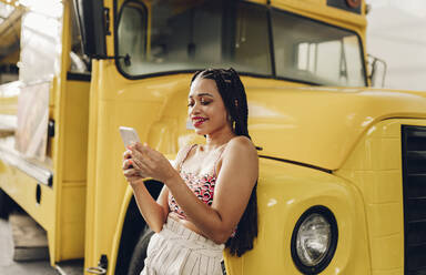Glückliche junge Frau mit Smartphone, die sich an einen Imbisswagen lehnt - JCCMF07293