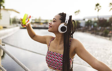 Smiling woman holding electric hand fan listening to music through wireless headphones - JCCMF07305