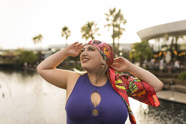 Happy young woman wearing colorful headscarf in front of lake - JCCMF07309