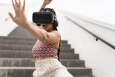 Junge Frau mit Virtual-Reality-Simulator gestikuliert auf einer Treppe - JCCMF07318