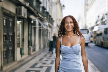 Happy woman with curly hair standing on footpath - DCRF01391