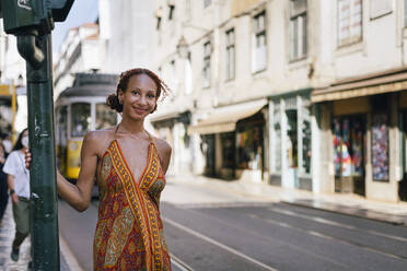 Smiling young woman standing by pole on footpath - DCRF01411