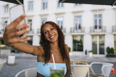 Happy woman taking selfie through smart phone sitting at sidewalk cafe - DCRF01432