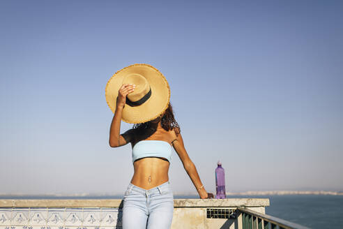 Junge Frau, die ihr Gesicht mit einem Hut bedeckt, steht an einer Mauer an einem sonnigen Tag - DCRF01459
