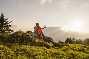 Smiling mother with daughter taking selfie through mobile phone sitting on rock - DIGF18784