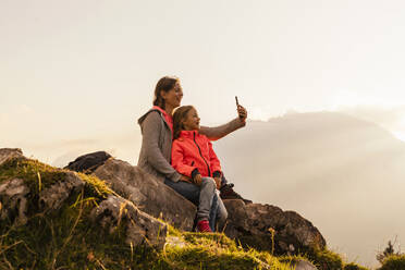 Glückliche Mutter mit Tochter, die ein Selfie mit ihrem Handy macht und auf einem Felsen sitzt - DIGF18785