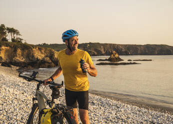 Happy man with bicycle holding water bottle at beach - UUF27230