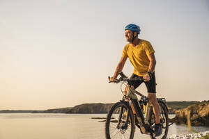 Älterer Mann fährt bei Sonnenuntergang Fahrrad am Ufer - UUF27232