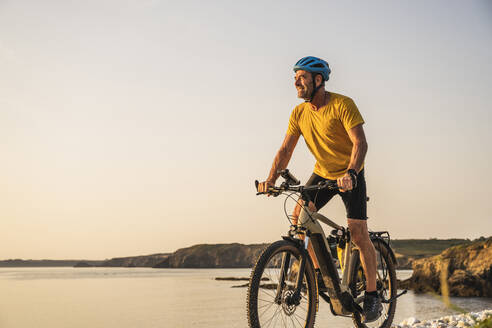 Älterer Mann fährt bei Sonnenuntergang Fahrrad am Ufer - UUF27232