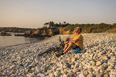 Älterer Mann ruht sich mit Fahrrad am Ufer bei Sonnenuntergang aus - UUF27235