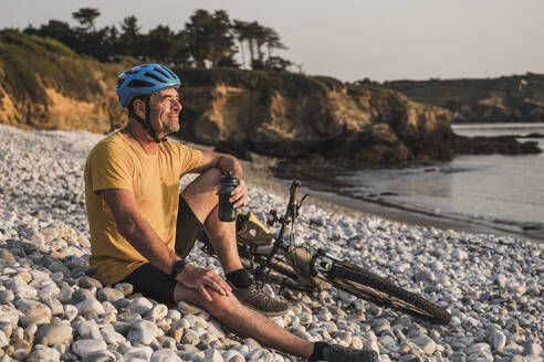 Thoughtful mature man sitting with bicycle on pebbles - UUF27240