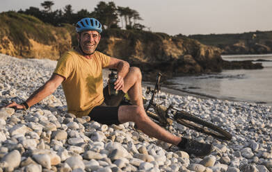 Glücklicher Mann sitzt am Fahrrad und hält eine Wasserflasche am Strand - UUF27242