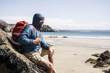Nachdenklicher reifer Mann mit Rucksack, der sich an einen Felsen am Strand lehnt - UUF27245