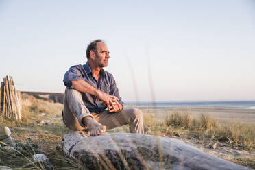 Thoughtful mature man sitting on log at sunset - UUF27269