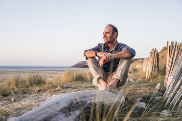 Smiling mature man sitting on log at beach - UUF27270