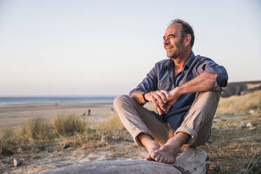 Smiling man sitting on log at beach - UUF27271